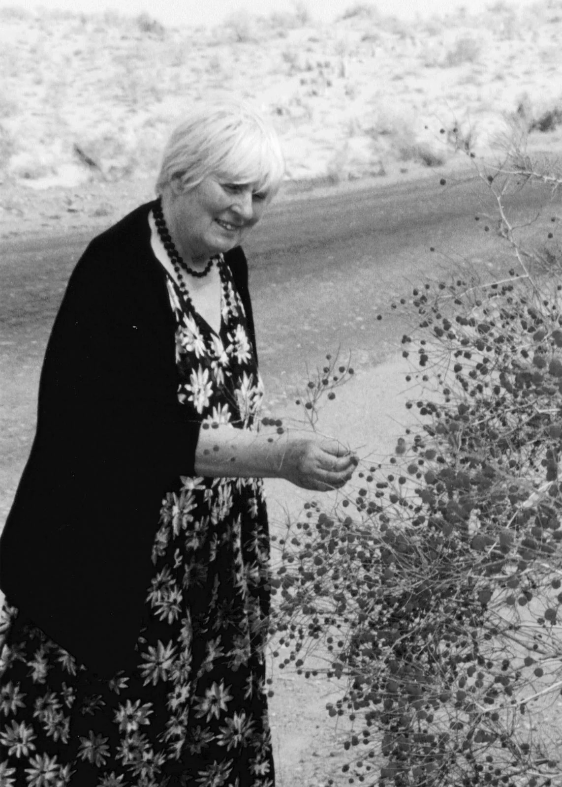 Black-and-white photograph of Sally smiling among wild plants, touching a delicate stem.